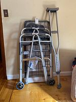 Two walkers, crutches, and folded shopping cart against a wall on hardwood floor