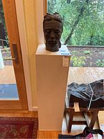 Frontal view of dark brown ceramic male bust on white pedestal near window with greenery outside