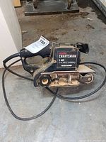 Side view of black Craftsman 1 HP belt sander on a concrete floor with power cord coiled, showing label and sanding belt wheels.
