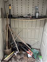 Photo of assorted garden tools including shovels, rakes, a broom, pruning shears, and a spray bottle in a storage shed.