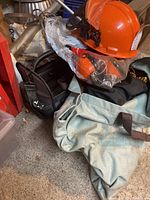 Photo showing orange chainsaw helmet with mesh eye protection and ear protection placed on top of black and light blue tool bags on concrete floor amid workshop clutter.