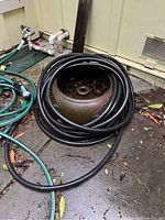 Black rubber garden hose coiled around a large round ceramic holder on outdoor concrete patio, showing the hose and ceramic holder and their condition.