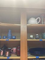 Wooden shelves holding a white teapot, some blue and black plates, various cups, and metal coffee filters, arranged with a mix of kitchenware items.