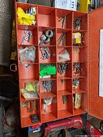 Orange steel box with 24 compartments filled with assorted screws, nuts, bolts, washers, and small parts, some in plastic bags.