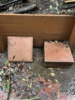 Five reddish-brown square concrete paver stones stacked on a wet ground near wooden structure, showing dirt and leaves.
