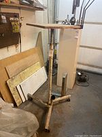 Photo showing worn wooden rectangular top with metal adjustable base, cross-shaped with wheels, standing in workshop area with plywood and tools.