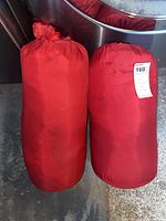 Two red sleeping bags inside red storage sacks, placed side by side on concrete ground. Show overall appearance and dimensions.