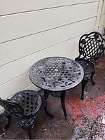 Photo showing round metal outdoor table and two matching chairs with ornate floral and lattice design, black with weathered silver accent.