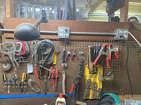 Wide view of mounted wooden pegboard holding various hand tools including screwdrivers, brushes, clamps, pliers, a lamp, and power outlet modules.