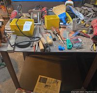 Metal work table cluttered with various hand tools, gloves, yellow storage bins, and hardware items.