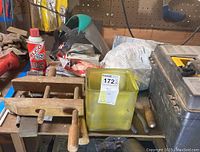 Table covered with various woodworking tools and supplies, including wooden bar clamps, a can of Elco 1620 spray adhesive, a yellow parts container, and a black toolbox.