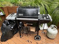 Front view of black Weber gas grill showing temperature gauge on lid, two control knobs, side shelves with grilling tools, propane tank to right, and grill cover on left side.