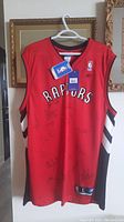Front view of the red Toronto Raptors jersey hanging on a hanger with visible NBA and Reebok tags and various signatures on the front.
