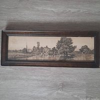 Full view of the framed antique etching/engraving artwork showing a rural landscape with buildings, water, and trees under natural lighting on a floor surface.