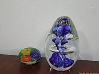 Two glass paperweights on a flat surface, smaller paperweight with colorful flowers and larger paperweight with blue bud-like abstract inside clear glass
