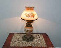 Lamp lit on a wooden table with lace doily, showing front floral glass shade and base.
