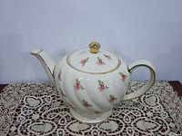 Front and side view of large white porcelain Sadler teapot with pink rose pattern and gold accents on lid knob, spout, and handle. Sitting on lace cloth.