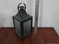 Front and side view of the metal and glass lantern placed on a wooden surface against a white wall, next to a ruler showing height.