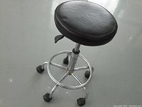 Photo showing the entire shop stool with black padded round seat, chrome base, and caster wheels on grey floor.