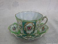 Front-side view of the small thin porcelain teacup placed on its matching saucer showing floral panels with gold trim and scalloped edges.