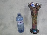 Full front view of the iridescent carnival glass vase next to a water bottle for scale, showing the trumpet shape and ruffled top.