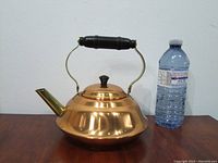 Front and side view of a vintage copper kettle with brass spout and handle, next to a water bottle for size reference.