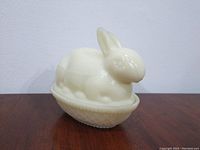 Opaque cream-colored glass bunny-shaped covered dish resting on wooden surface, showing full exterior view.