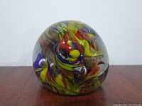 Front view of the round glass paperweight showing its colorful swirl pattern inside on a wooden surface against a white wall background.