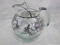 Side view of vintage glass pitcher with silver overlay motifs over the body and rim, showing round shape and handle.