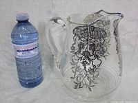 Wide shot showing the clear glass pitcher next to a water bottle for size comparison, highlighting the silver floral overlay decoration on the upper half and spout of the pitcher.