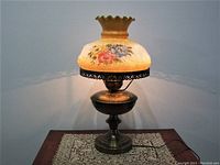 Frontal view of the vintage brass hurricane style electrified table lamp on a lace doily on a wooden surface against a white wall. Shows the full lamp with its base and floral hand-painted glass shade lit to display warm light.