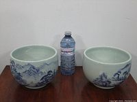 Two large ceramic bowls side by side with a water bottle placed between them for size reference on a wooden surface against a white wall background. Bowls have hand-painted blue landscape designs depicting mountains and trees.