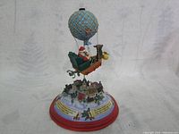 Full view of the vintage tin wind-up music toy showing Santa in a hot air balloon above a snowy village with detailed miniature houses, trees, and a decorated base.