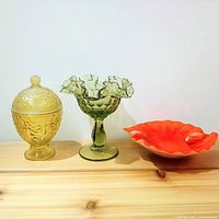 Front view of three glass items: yellow covered dish, green hobnail pedestal bowl, orange ruffled edge bowl.