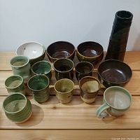 Overview of entire lot showing 10 cups, 4 bowls, and 1 tall vase on wooden surface, various sizes and glazes visible