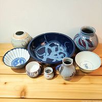 Overview of seven vintage Asian ceramic items showing bowls, jars, and vases in blue, white, red, and beige tones arranged on wooden surface.