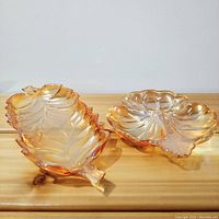 Two Indiana Glass lusterware dishes, leaf and shamrock shaped, with peach-gold iridescent finish displayed on wooden surface.