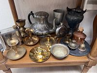 Collection of mixed metal candle holders, teapot, flask and other decorative metal items on wooden shelf