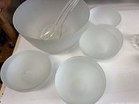 Frosted glass salad serving bowl with multiple smaller matching bowls and clear plastic salad servers visible on a white surface.