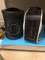 Two Honeywell personal small space heaters sitting side by side on floor: one cylindrical with dual dials, one tower style.