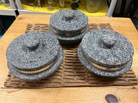 Set of three Korean stone pots with lids shown from above and front angles on a wooden surface, showing textured gray stone, round shape, and metal band.