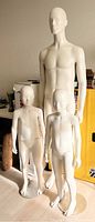 Full view showing adult male mannequin and two child mannequins standing side by side on floor with light and shadow visible
