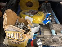 Close-up photo of open cardboard box containing various screws, partially torn box labeled 'NAILS PSS' with additional tools and hardware visible in background.