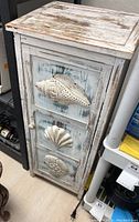 Close-up side-angle photo showing the three white metal nautical shell decorations mounted vertically on the cabinet door and the distressed paint finish on the edges.