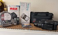 Photo showing Sony Handycam camcorder, RCA BAMM video tape recorder, desktop cassette recycler in box, remote control, cables, and carrying bags arranged on a shelf.