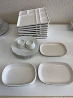 Photo showing stacks of white ceramic plates with various shapes including square divided plates and oval plates, plus two small white salt and pepper shakers.