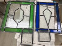 Two stained glass panels side by side, one with green border and diamond shaped center, and one with blue border and kite shaped center, showing full panels and clear visibility of glass details.
