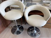 Front angle showing two white leatherette stools with round cushions, open backrest, chrome pedestal base, and footrest ring.