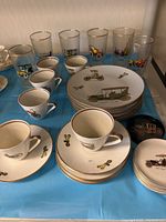 Overview of porcelain cups, saucers, plates, and drinking glasses with vintage car designs and gold rims.