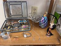 Full view of the trinket box with pressed flower pewter lid open showing assorted vintage jewelry, pins, a watch, and silver-tone bracelets on a wooden surface alongside the glass swan dish, perfume bottle atomizer, and a small decorative item.